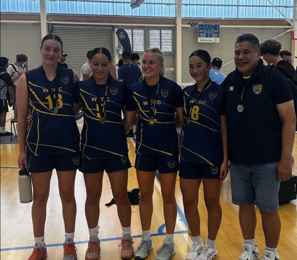 Zara Thomas, far left in a number 13 WEGC singlet, with four teammates and a coach wearing medals courtside after a tournament.